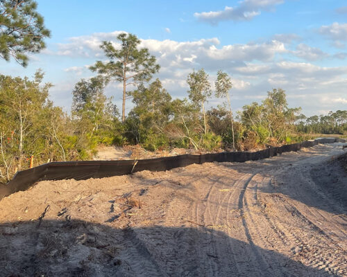 Dirt path with tire tracks beside black silt fence, trees nearby.