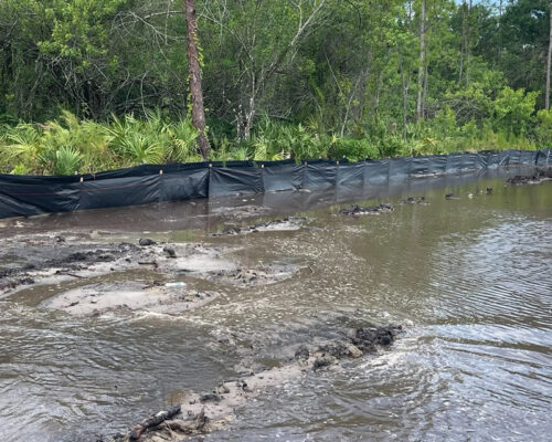 Flooded mud by a black silt fence, trees behind.