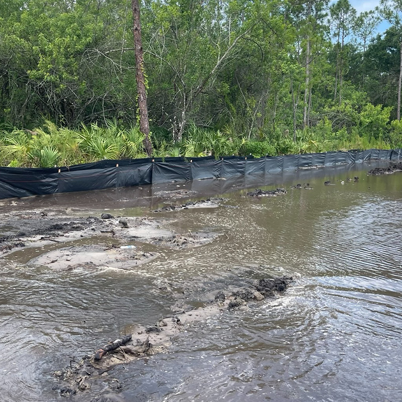 Flooded mud by a black silt fence, trees behind.