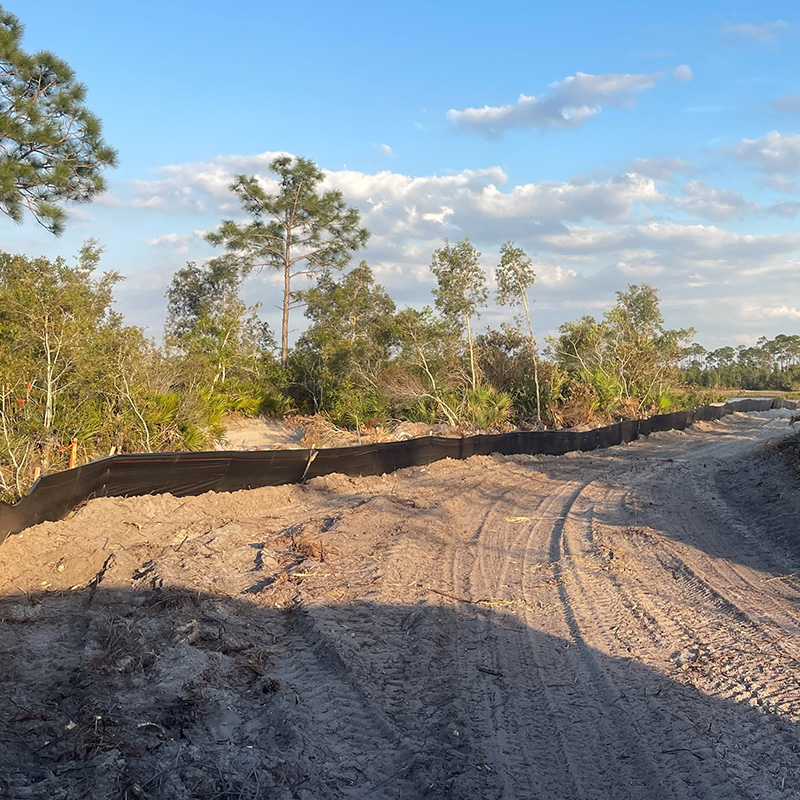 Dirt path with tire tracks beside black silt fence, trees nearby.