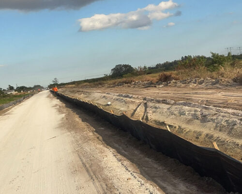 Dirt road by construction, black silt fence, two workers visible.