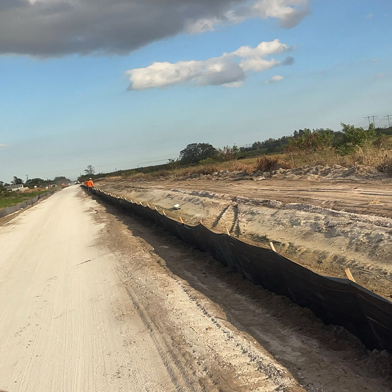 Dirt road by construction, black silt fence, two workers visible.