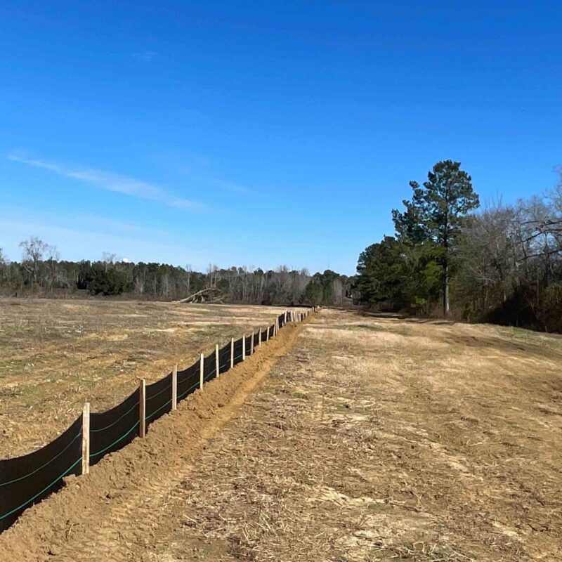 Silt fence in grassy field, trees and blue sky behind.