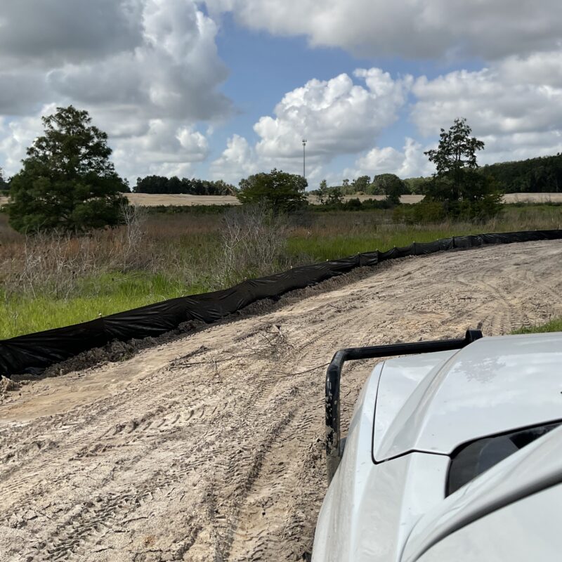 White vehicle on sandy road by black silt fence, fields, sky.
