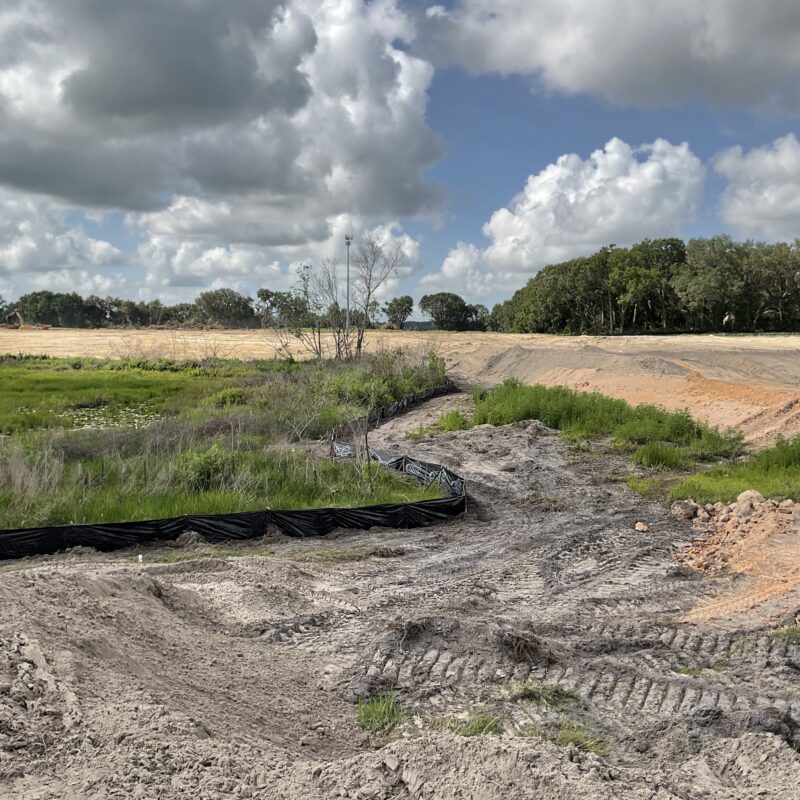 Construction site with dirt, tire tracks, grass, silt fence, trees.