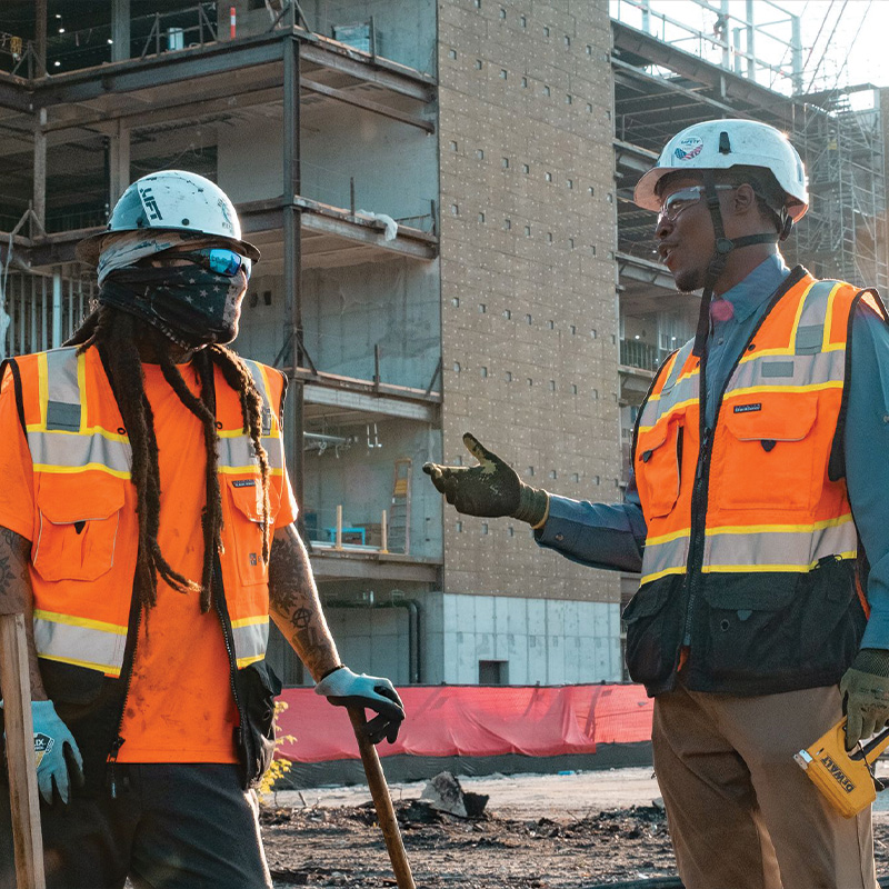 Two construction workers talk at a site with a half-built structure.