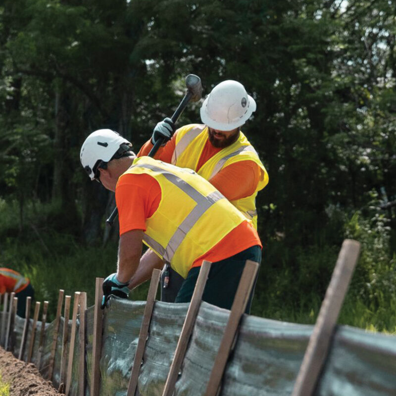 Two workers in safety gear install a silt fence along a trench.
