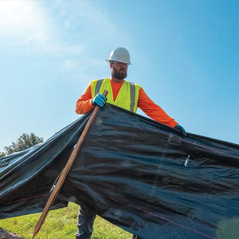 Worker installs black barrier on grass under blue sky.