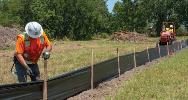 Worker installs stakes for silt fence; another drives utility vehicle.