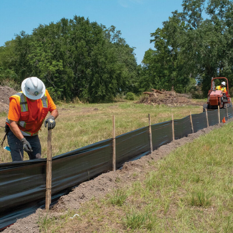 Worker installs stakes for silt fence; another drives utility vehicle.