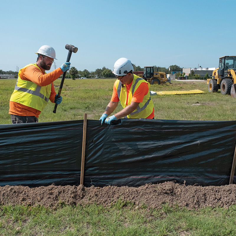 Workers install a black barrier with stakes; bulldozers behind.