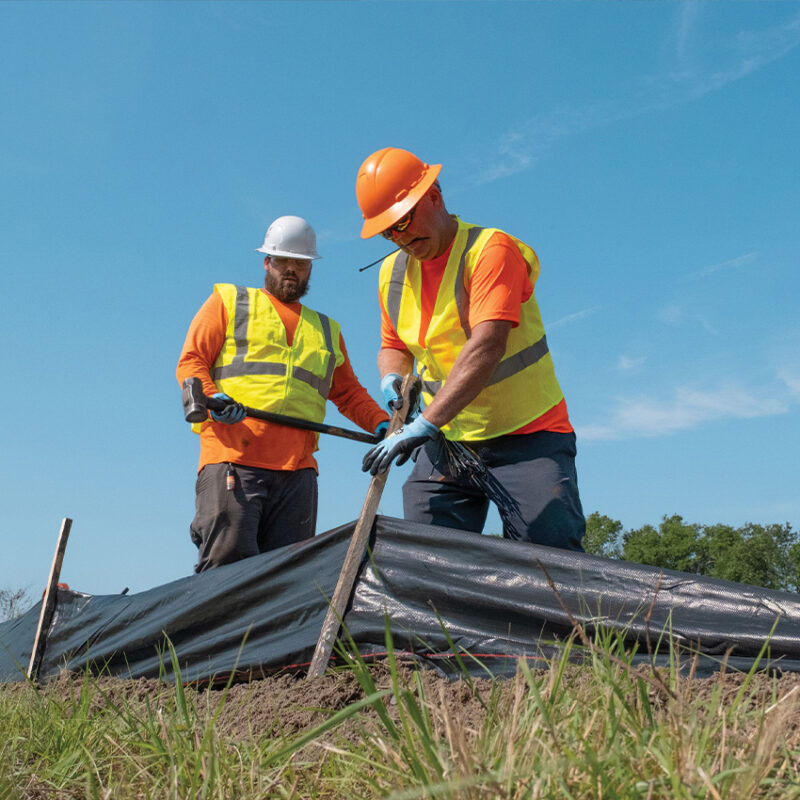 Workers in safety gear install black barrier by grassy area.