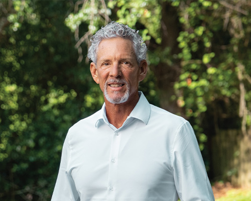 Older man with curly gray hair outdoors among trees.
