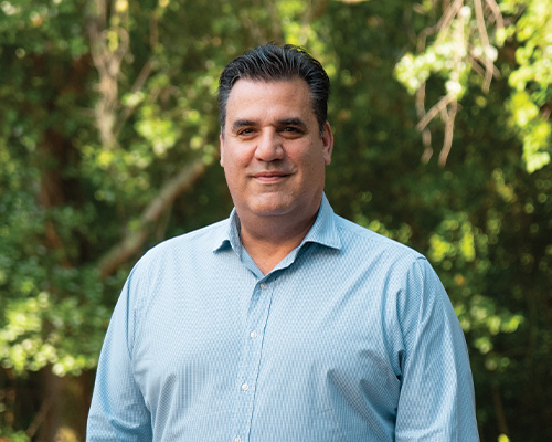 Man in light blue shirt stands outside with greenery behind.