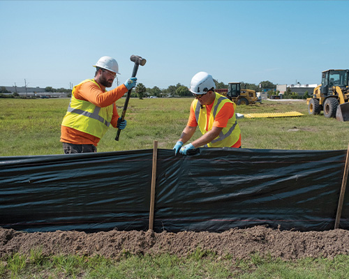 Two workers install a black silt fence outdoors.