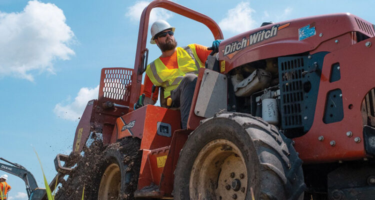Worker in vest and hard hat digs with red Ditch Witch.