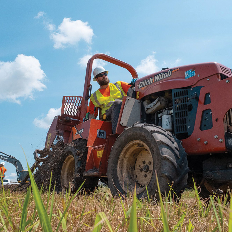Worker in vest and hard hat digs with red Ditch Witch.