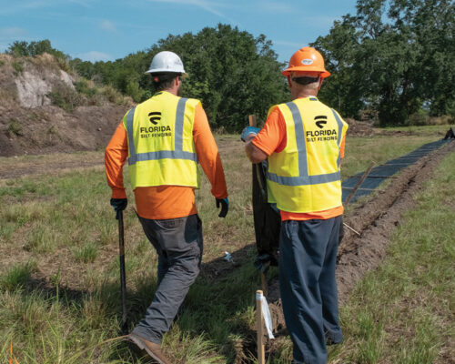 Two helmeted workers in vests by a trench in grassy field.