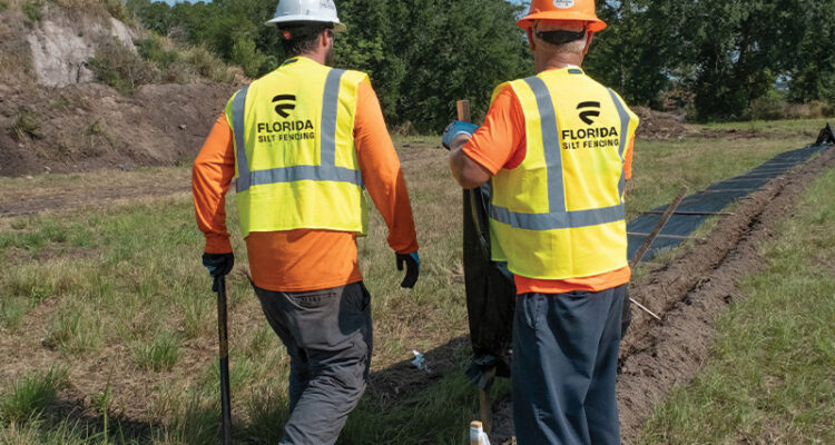 Two helmeted workers in vests by a trench in grassy field.