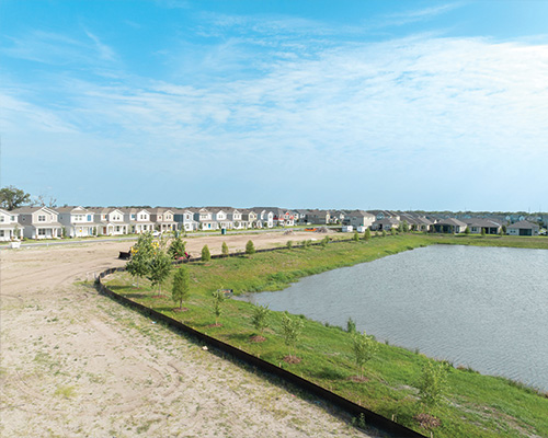 Suburban houses by a pond, dirt and young trees in front.
